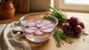 Ciotola di vetro con fette di cipolla rossa in ammollo in acqua su tavolo di legno, con cipolle e rosmarino